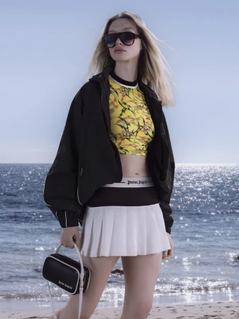Model wearing dark sunglasses, black jacket, yellow graphic top, and white pleated skirt on a sandy beach against the ocean.