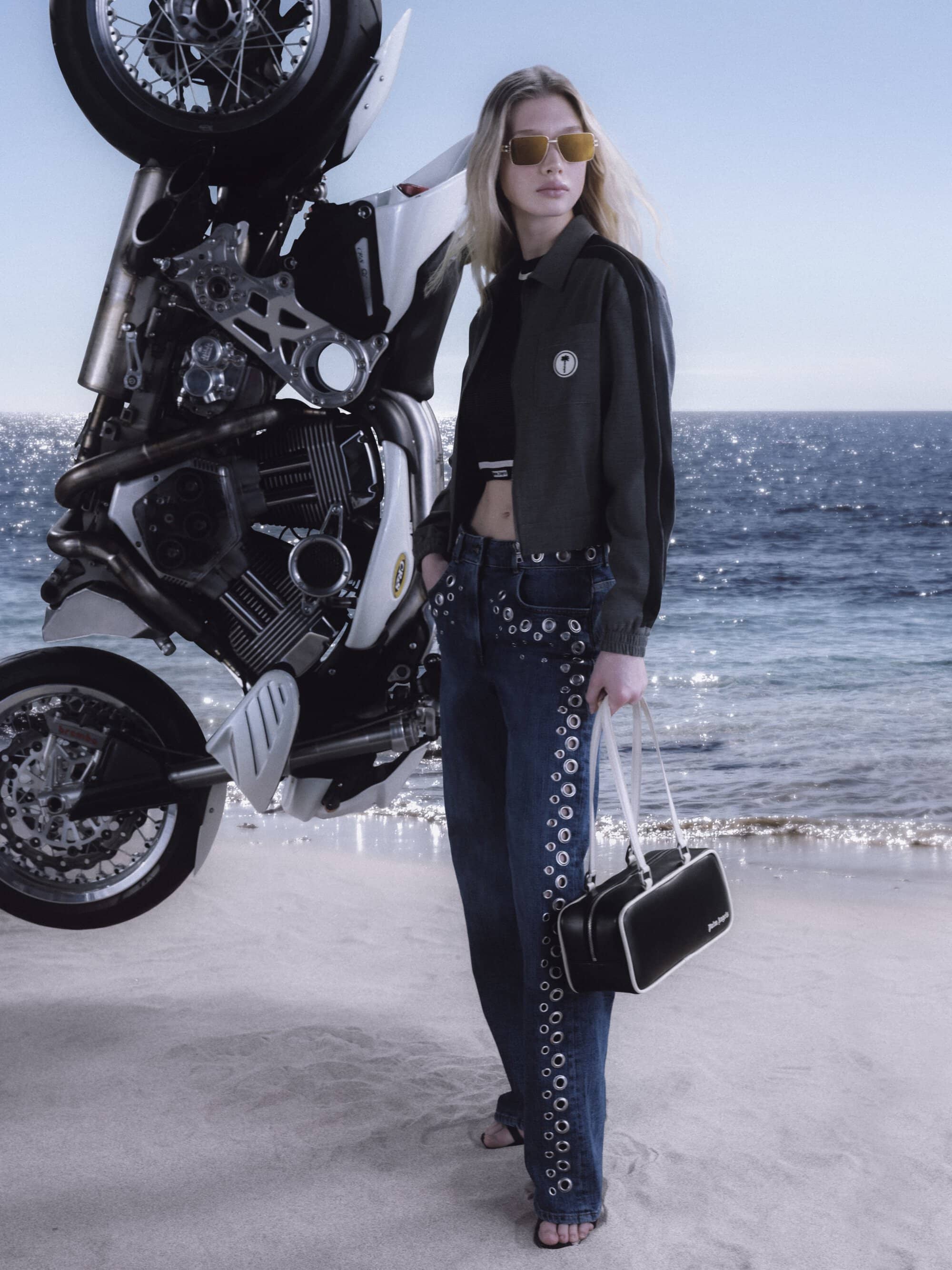 Woman in dark jacket and grommet jeans holding a black handbag standing beside a motorcycle on a sandy beach