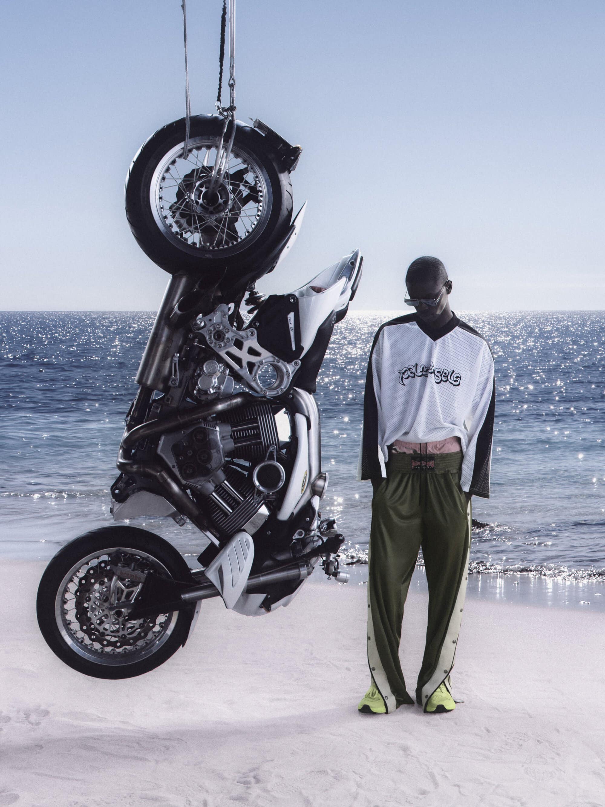 Black man standing on a sandy beach next to a suspended vertical motorcycle for Palm Angels ad campaign