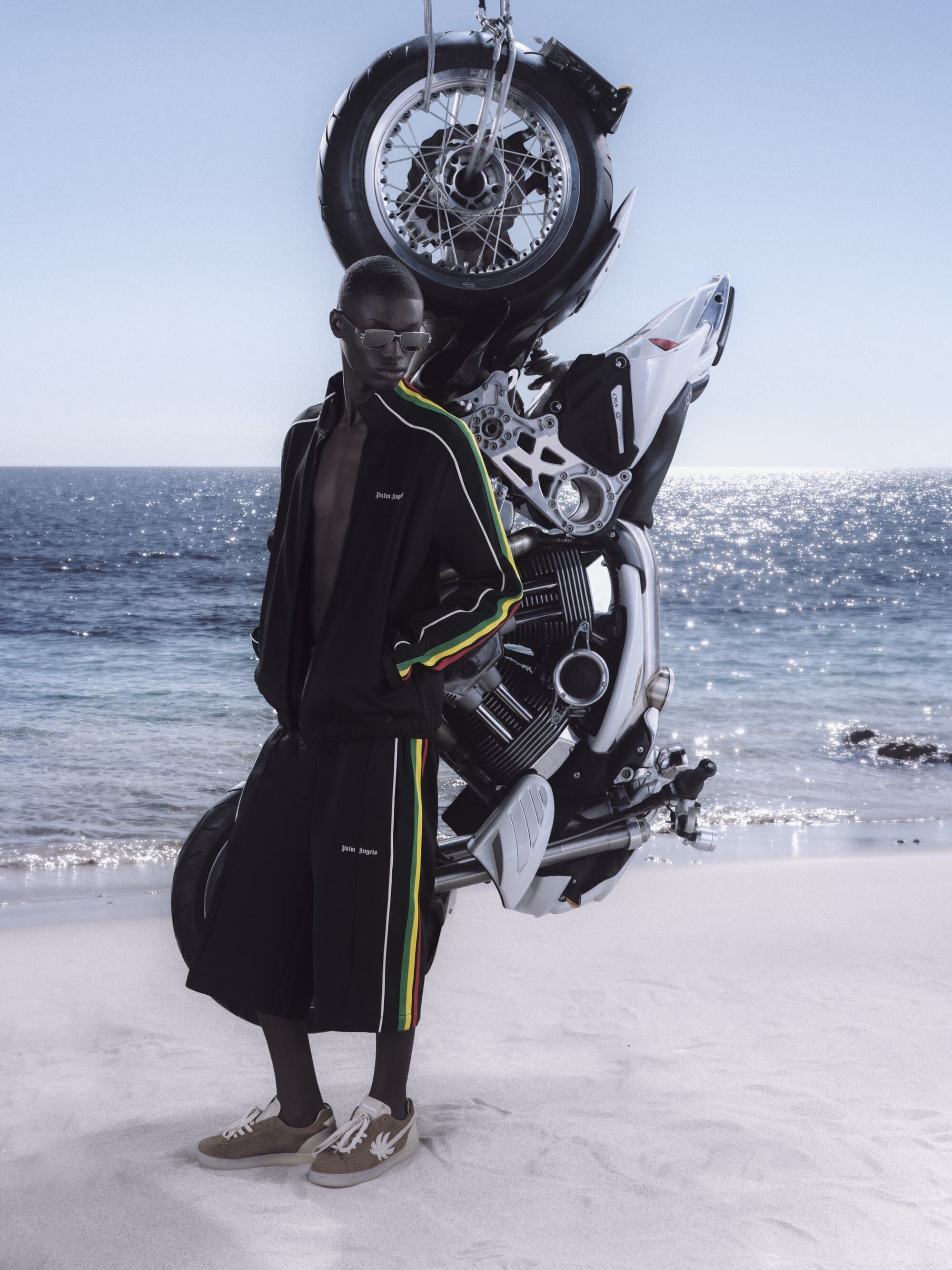 Model standing on a sandy beach next to a motorcycle hoisted in the air against the ocean for Palm Angels