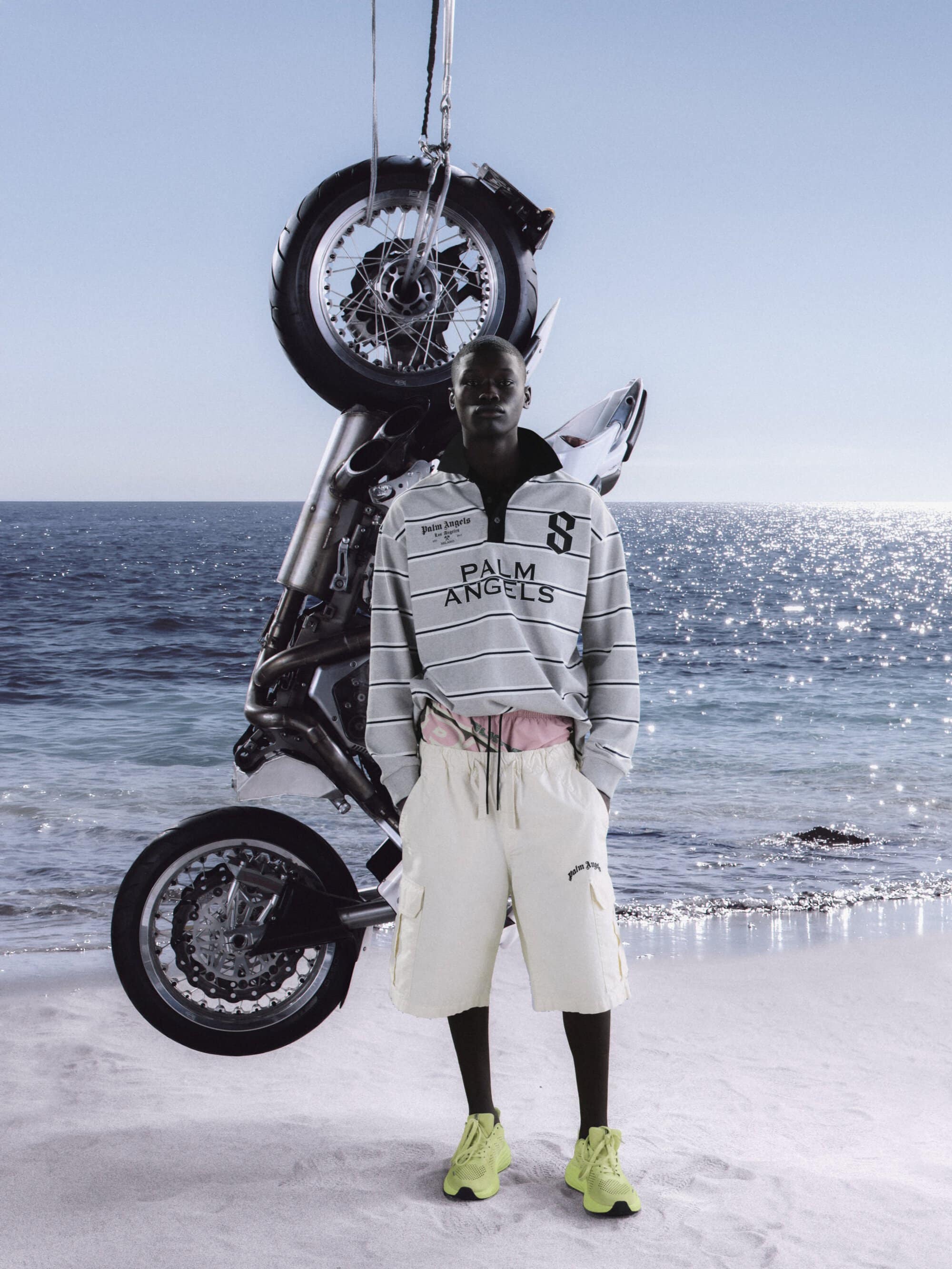 Male model in Palm Angels striped shirt and shorts poses on a sandy beach with a motorcycle suspended behind him on a sunny day.