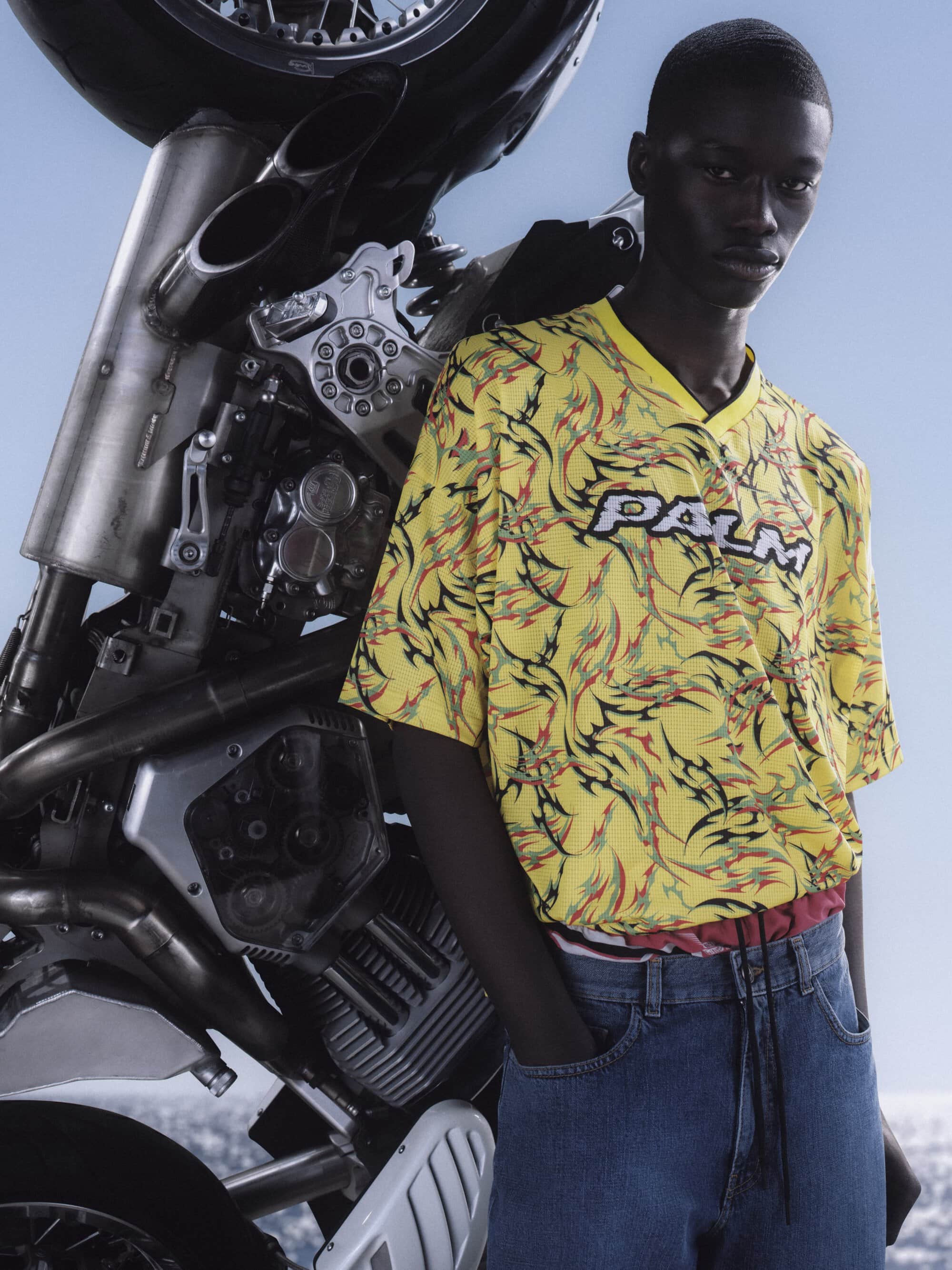 Black man wearing a yellow patterned t-shirt and blue jeans leaning against a motorcycle outdoors under a clear sky.