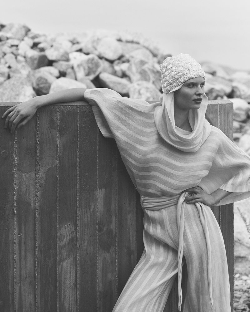 Monochrome fashion shot of a stylish woman wearing a striped jumpsuit and patterned hat, posing by a concrete structure with rocks behind.