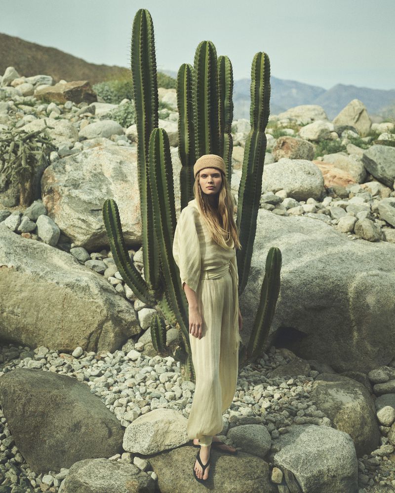 Model standing among tall cacti and rocky terrain wearing a light neutral jumpsuit and matching beanie.
