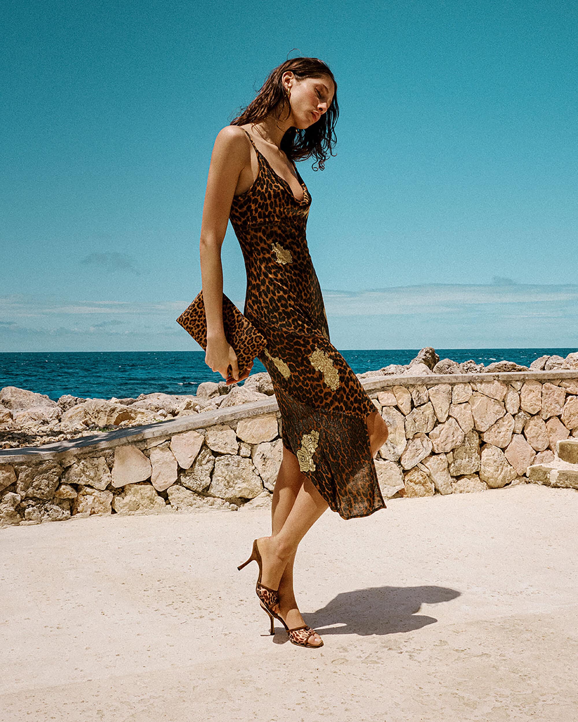 Model wearing a leopard print slip dress and matching heels holding a clutch by the ocean for Stuart Weitzman.