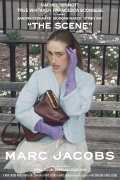 Model seated on a park bench wearing a light blue jacket and purple gloves holding a dark red leather handbag