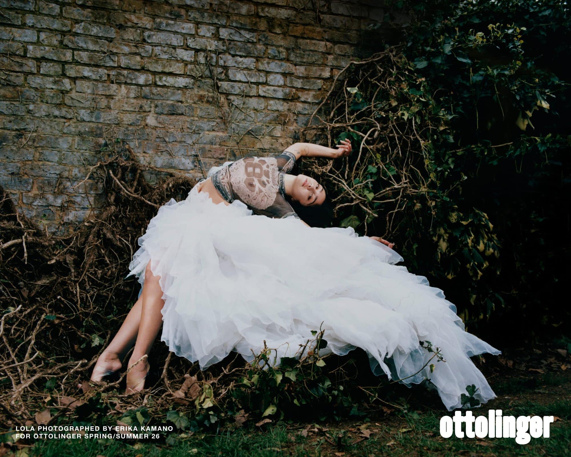 Woman in ethereal white gown poses against brick wall and greenery in a fashion editorial, exuding elegance and mystery in natural light.