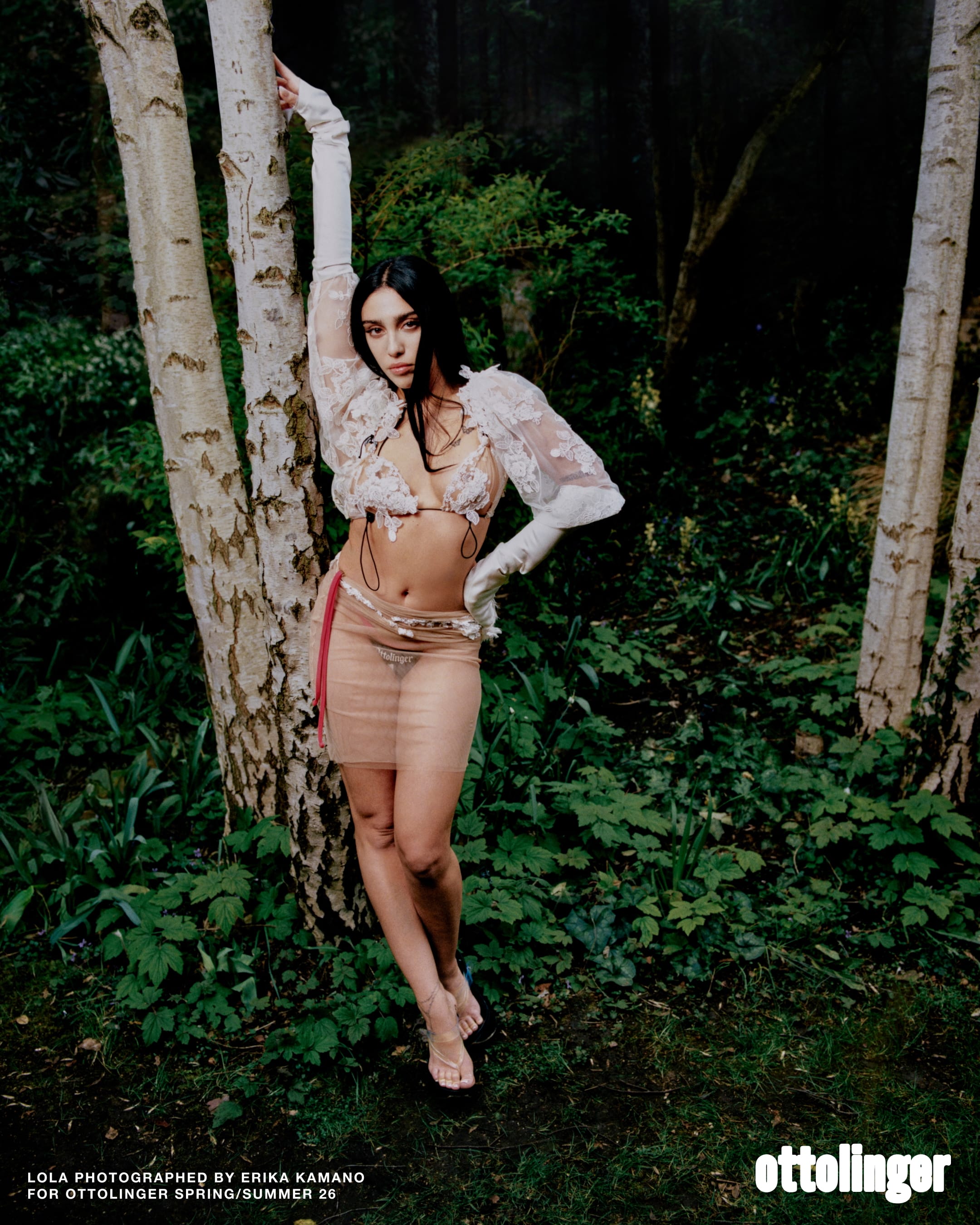 Model Lola Leon posing against a white birch tree in a lush green forest wearing a sheer lace top and skirt for Ottolinger.