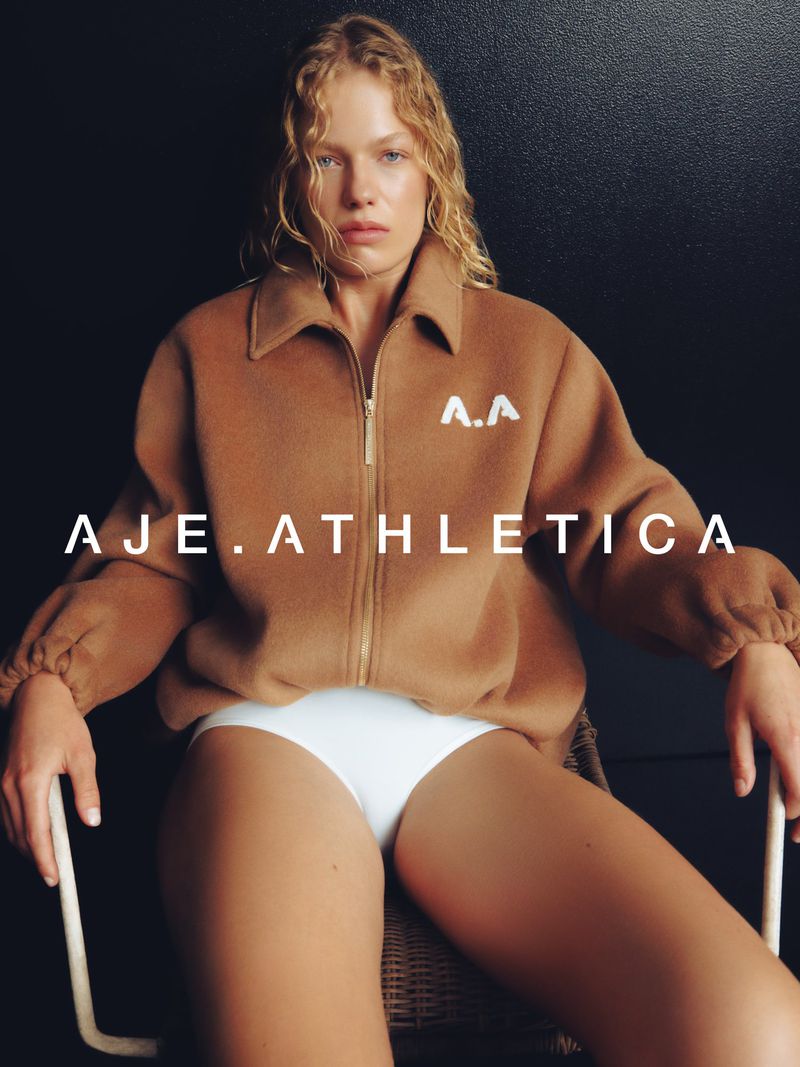 Model wearing a brown zip-up jacket sitting in a chair against a dark background for Aje Athletica Winter 2026