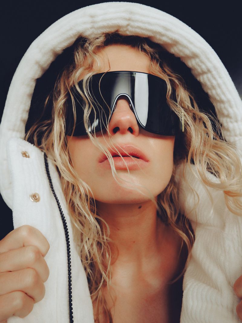 Close-up portrait of a blonde model wearing oversized black sunglasses and a white hooded puffer jacket