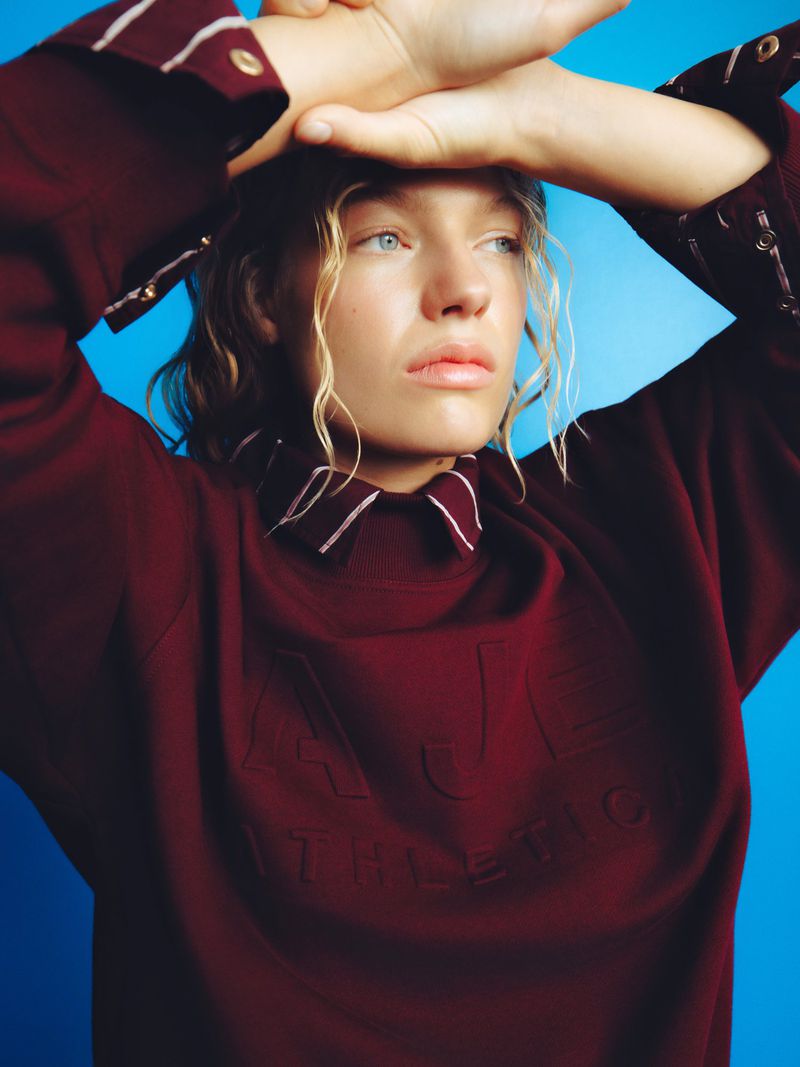 Model in a burgundy sweatshirt with arms raised against a bright blue background for Aje Athletica Winter 2026