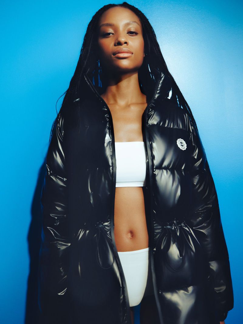 Model wearing a glossy black puffer jacket and white top against a vibrant blue studio backdrop