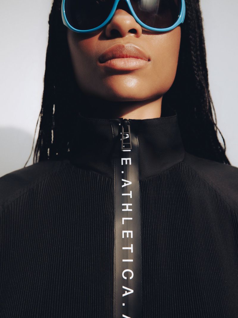 Close-up portrait of a model wearing bright blue sunglasses and a black zip-up top with branded taping against a neutral background.