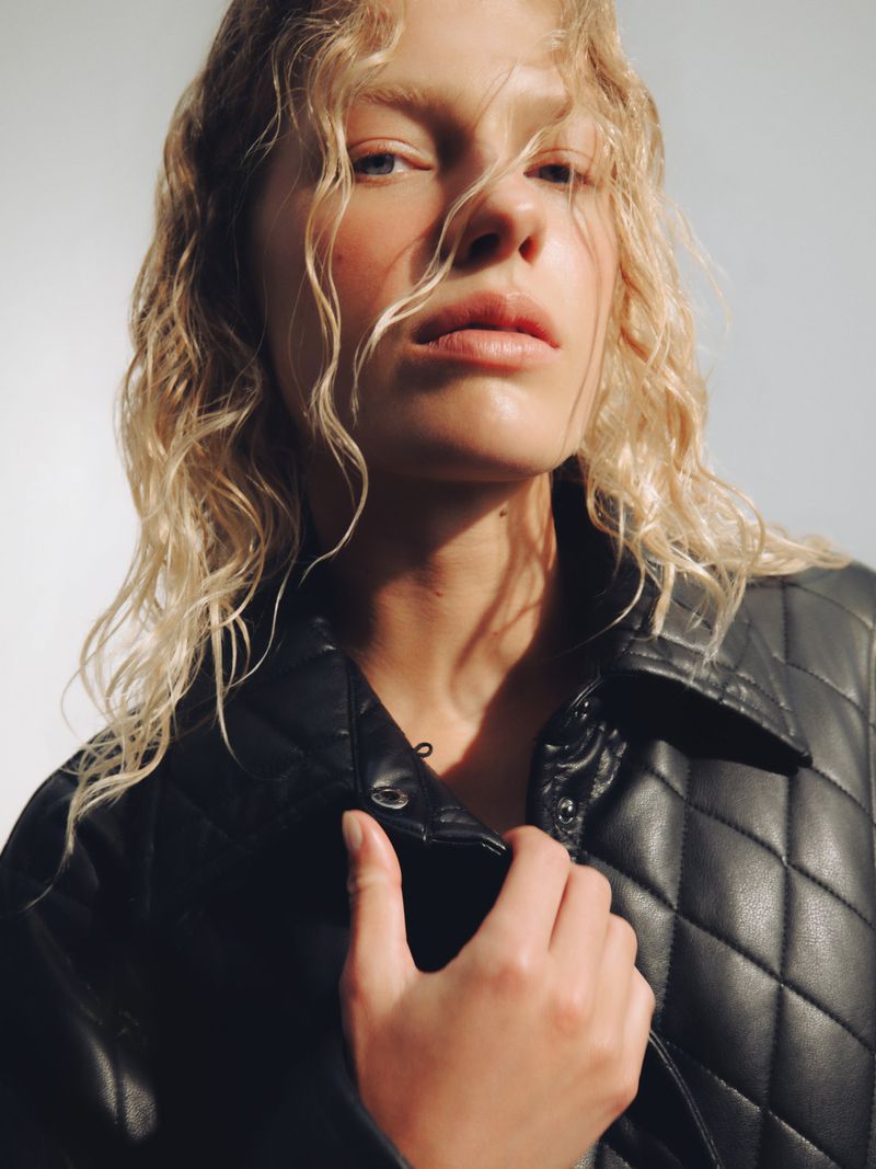 Close-up portrait of a blonde model wearing a black quilted jacket against a neutral backdrop.
