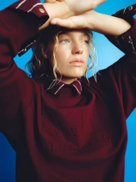 Model in a burgundy sweatshirt with arms raised against a bright blue background for Aje Athletica Winter 2026