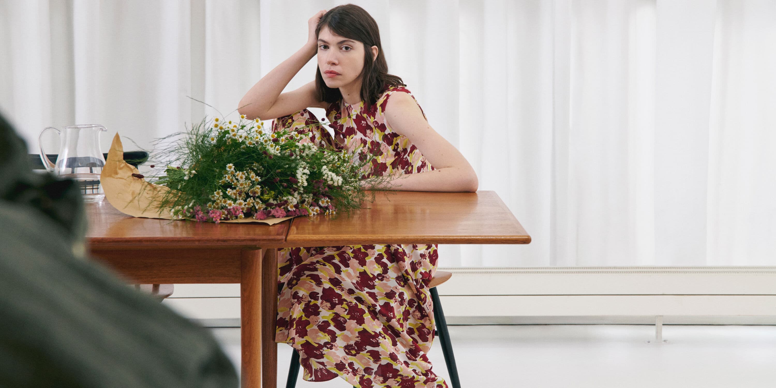 A woman wearing a floral dress seated at a wooden table next to a bouquet of wildflowers against a white curtain background.