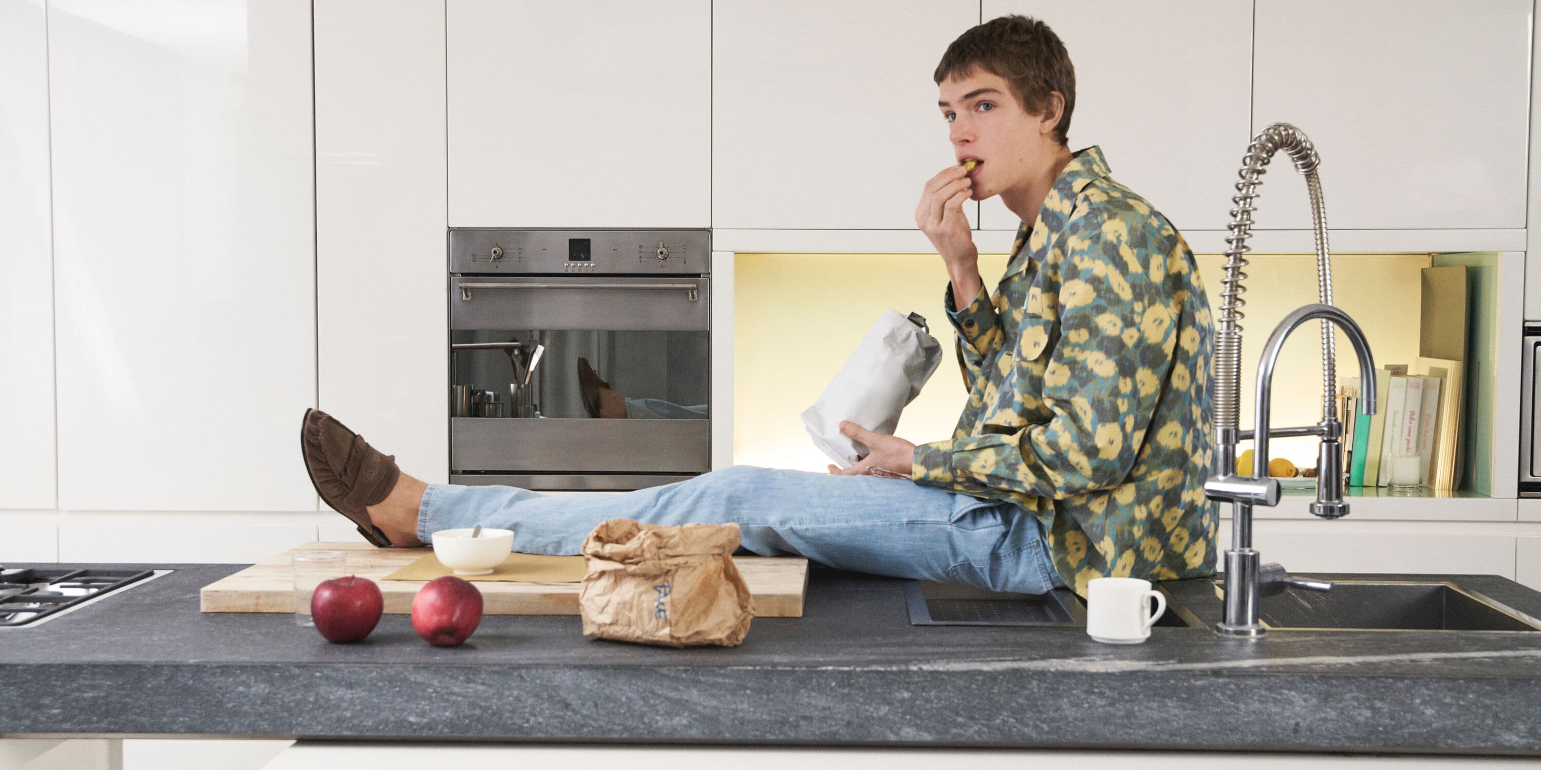 Young man in patterned shirt eating snack seated on modern kitchen counter