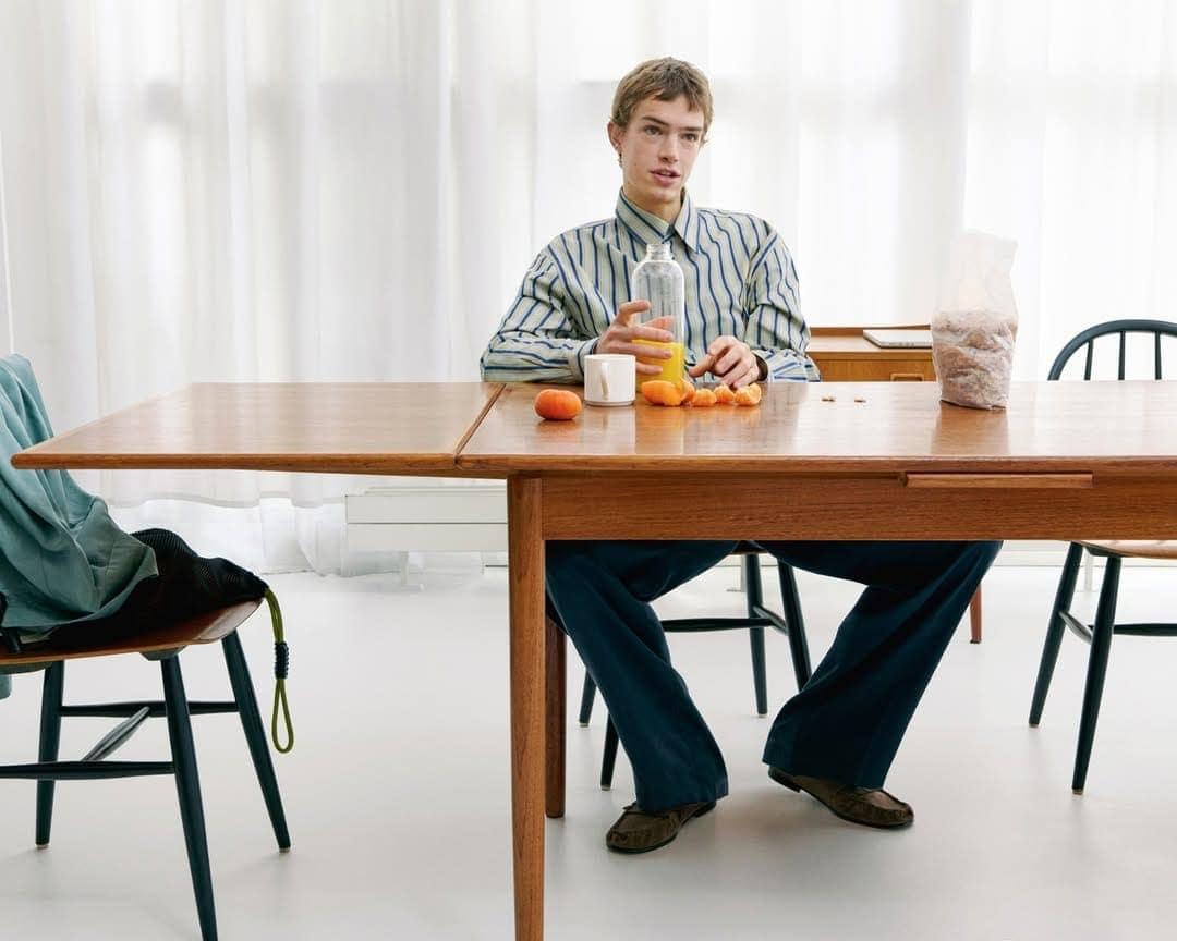 Young man in a striped button-down shirt seated at a wooden dining table holding a glass bottle with orange juice.