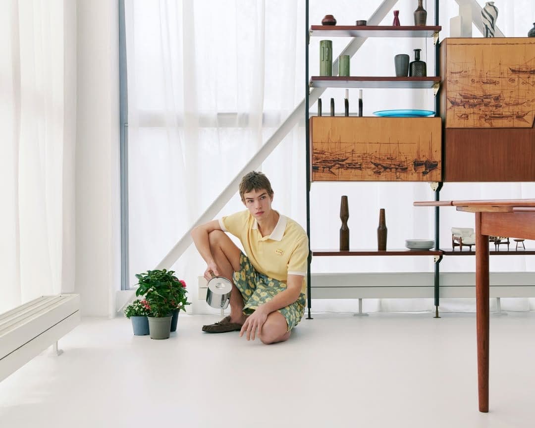 Young male model crouching beside potted plants holding a watering can in a bright interior setting.