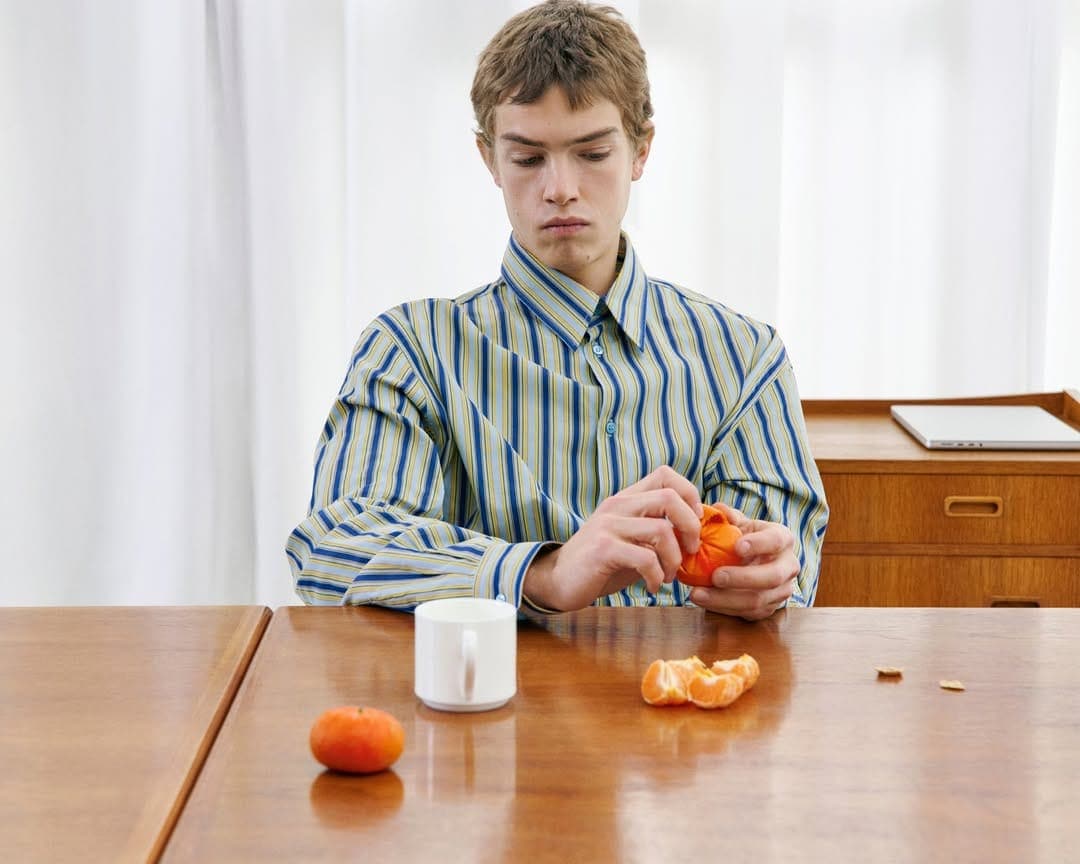 Man peels an orange at a wooden table with a white mug and laptop in the background, wearing a blue and yellow striped shirt.