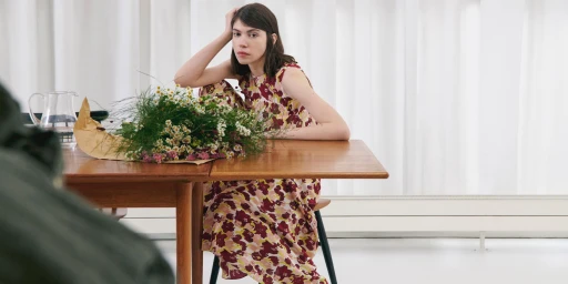 A woman wearing a floral dress seated at a wooden table next to a bouquet of wildflowers against a white curtain background.
