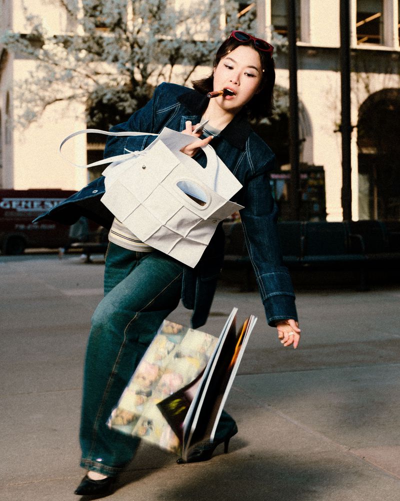 Woman in denim jacket and jeans drops white tote bag, papers, and magazine while walking on city street with blurred building.