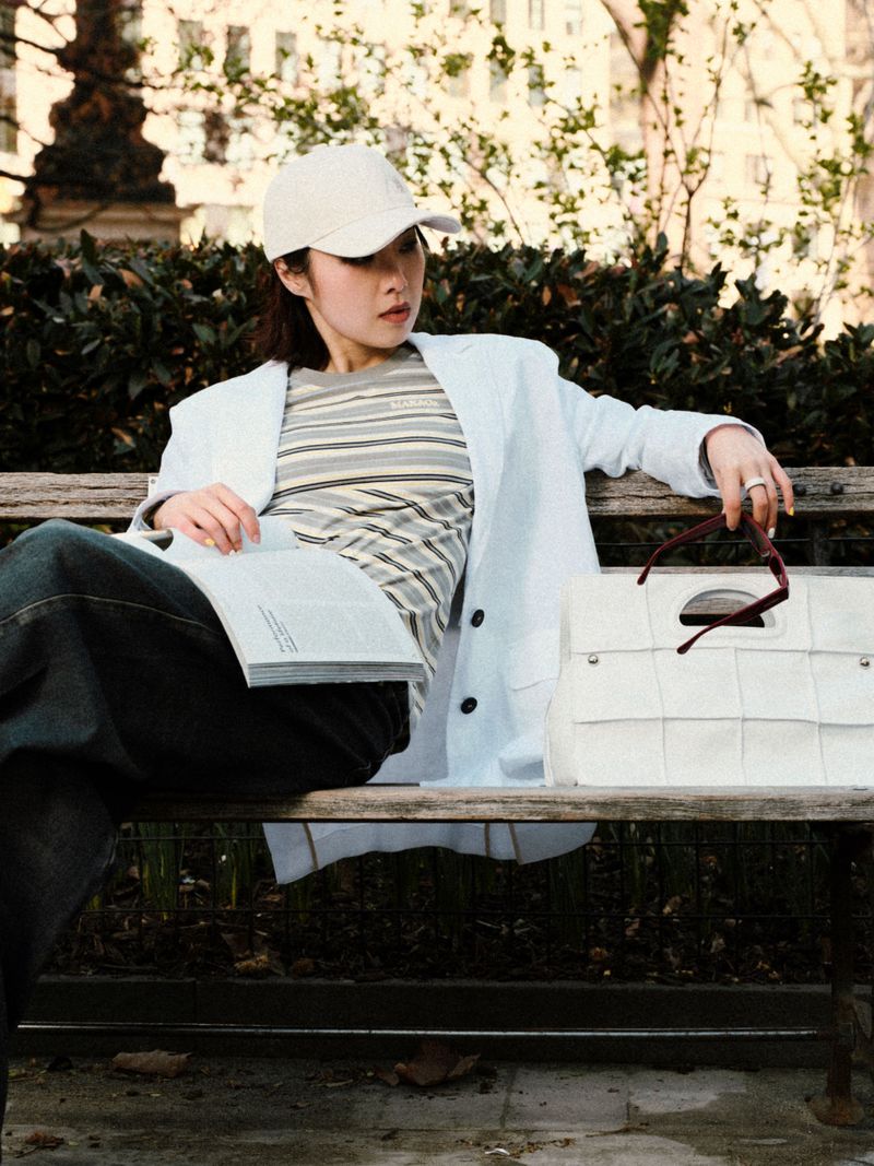 Woman wearing white cap and striped t-shirt seated on a wooden bench holding an open magazine with a white handbag beside her.