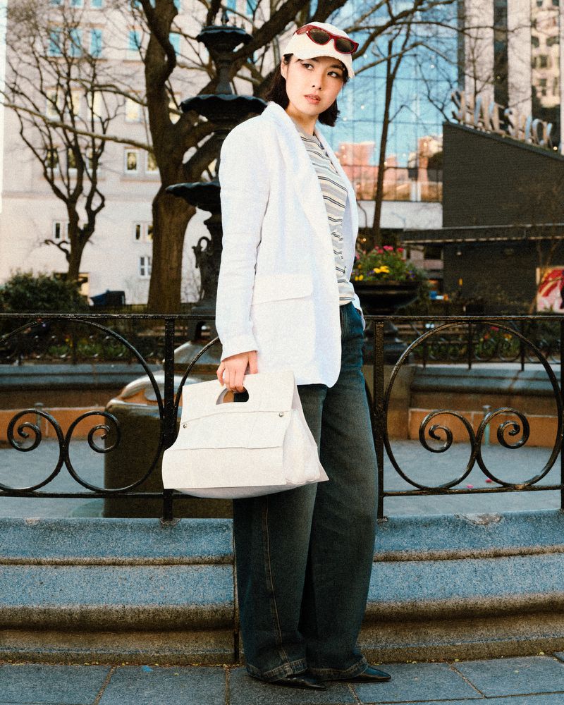 Model in white blazer and cap holding large handbag outdoors near iron railing