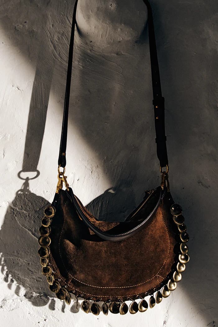 Brown suede shoulder bag with metallic embellishments hanging against a textured white wall.