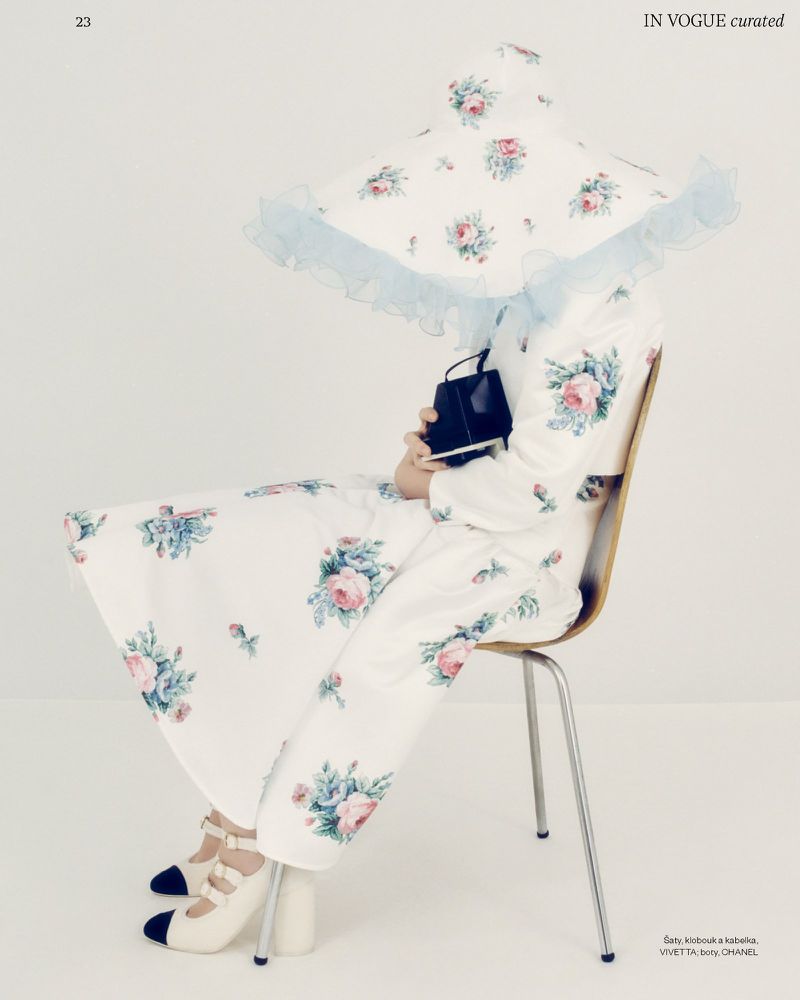 Woman in floral dress and hat sitting on chair holds black purse, posing in a light studio against a white seamless backdrop.