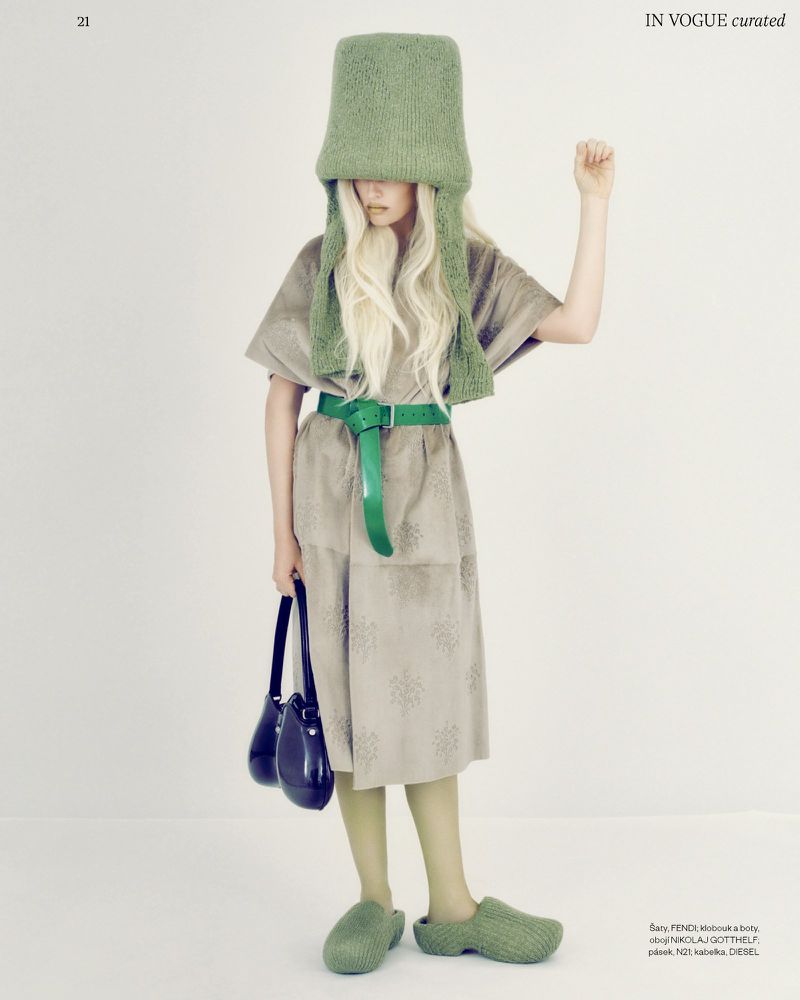 A person in an unusual green bucket hat and green accessories poses in a beige dress against a clean studio backdrop, holding a black bag.