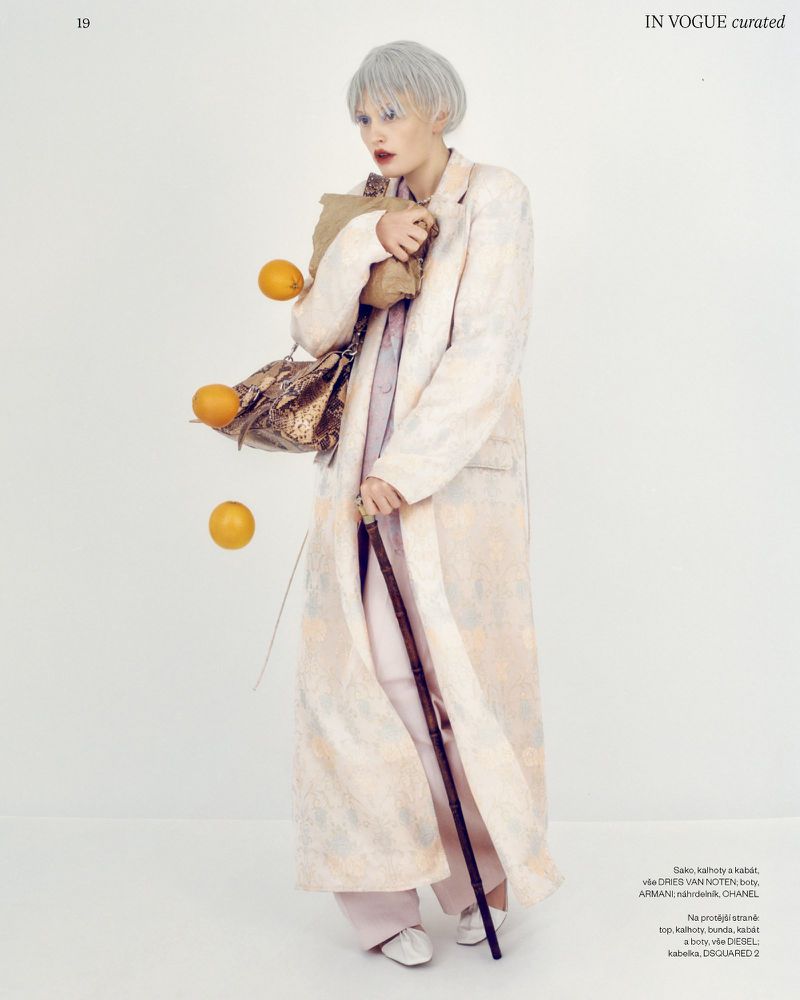 Fashion editorial shot of a woman in a long coat, carrying a brown paper bag and snakeskin purse, with oranges in a white studio.
