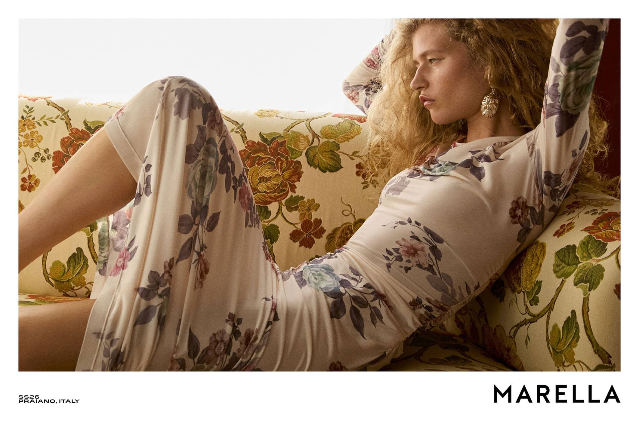 Model with long curly blonde hair wearing a floral dress reclining on a patterned sofa for Marella