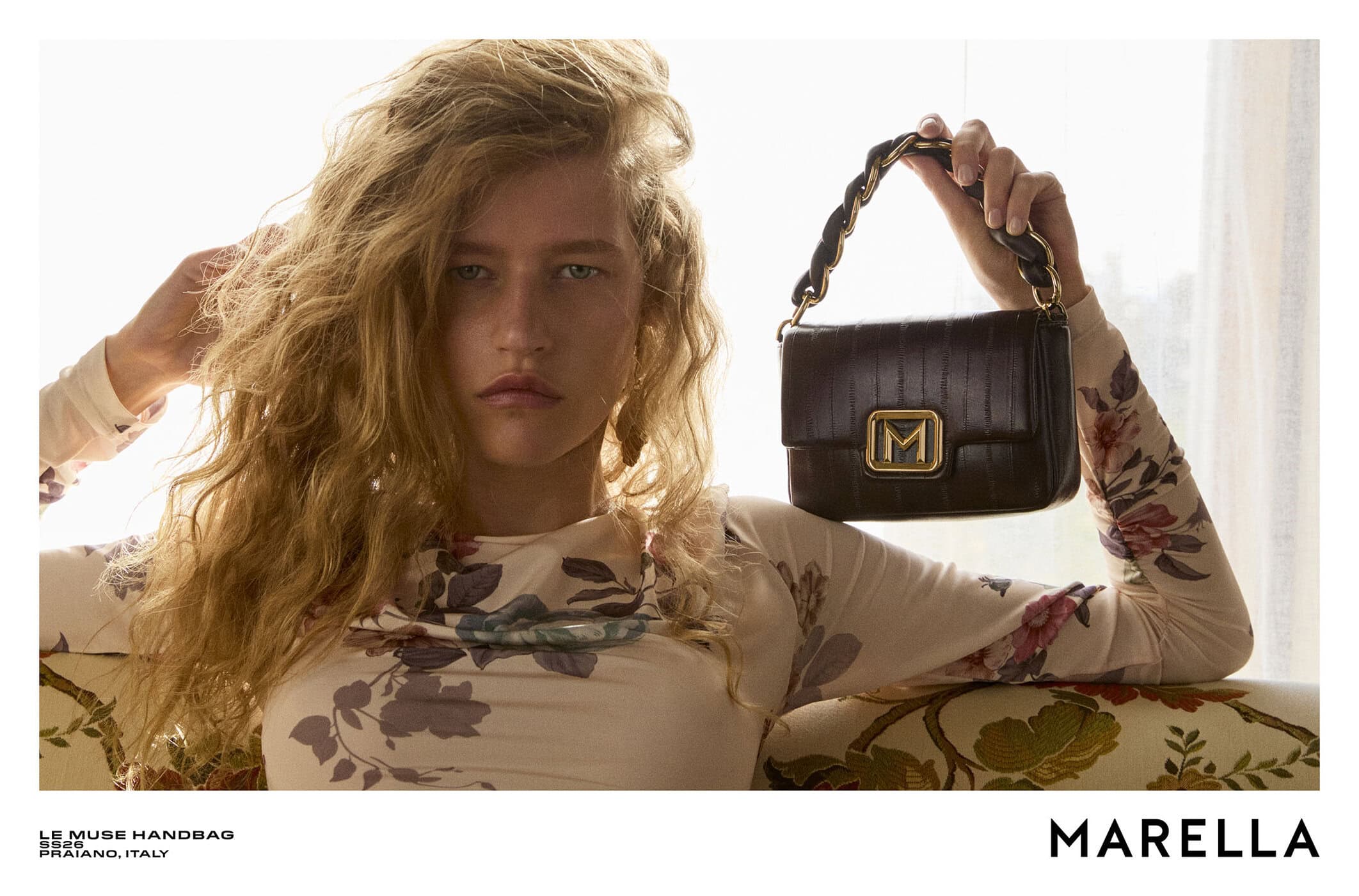 Model with long blonde hair holding a dark leather handbag in an editorial advertising campaign for Marella.