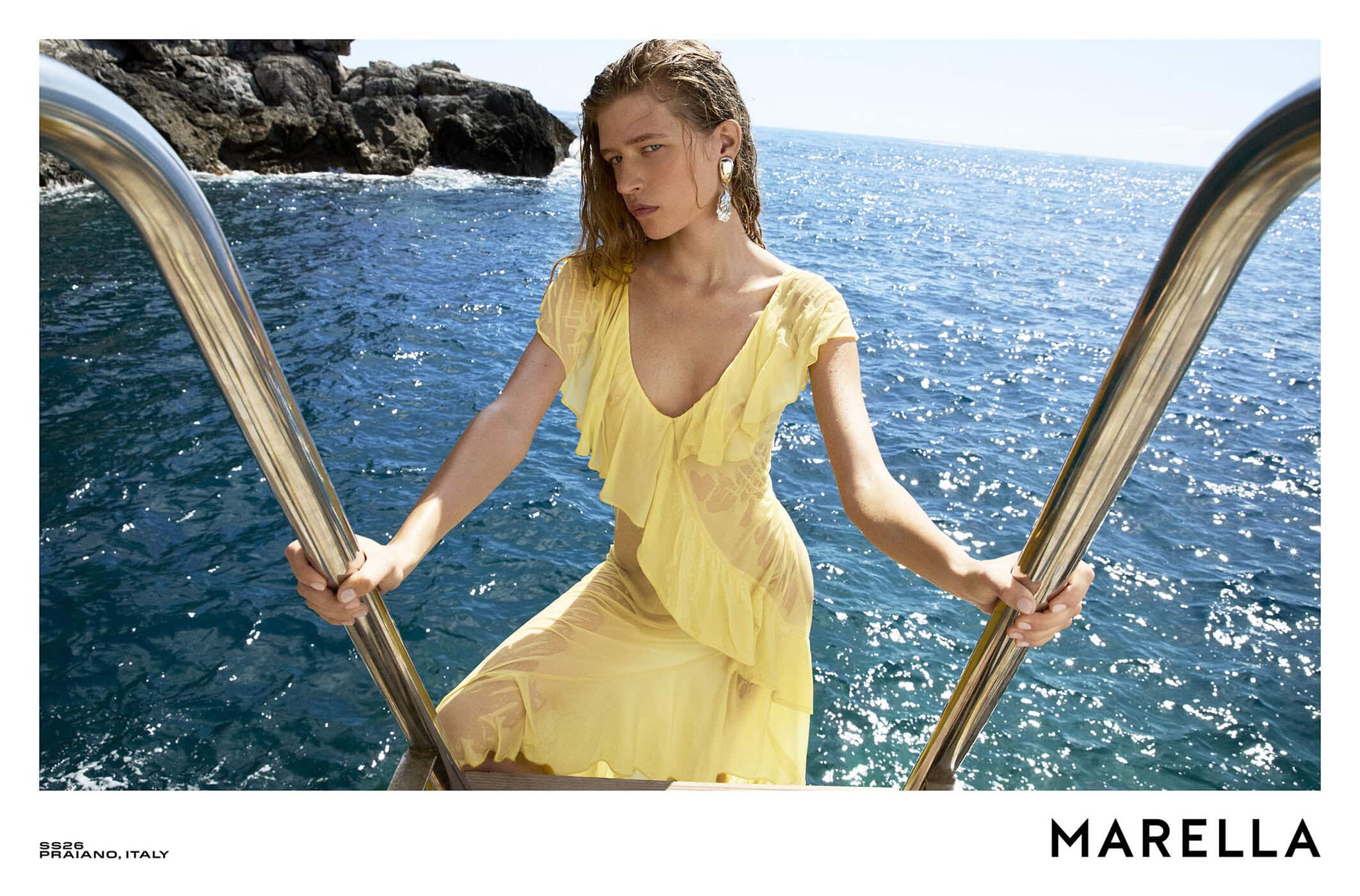 Model in a yellow ruffled dress holding metal pool rails by the sea for Marella