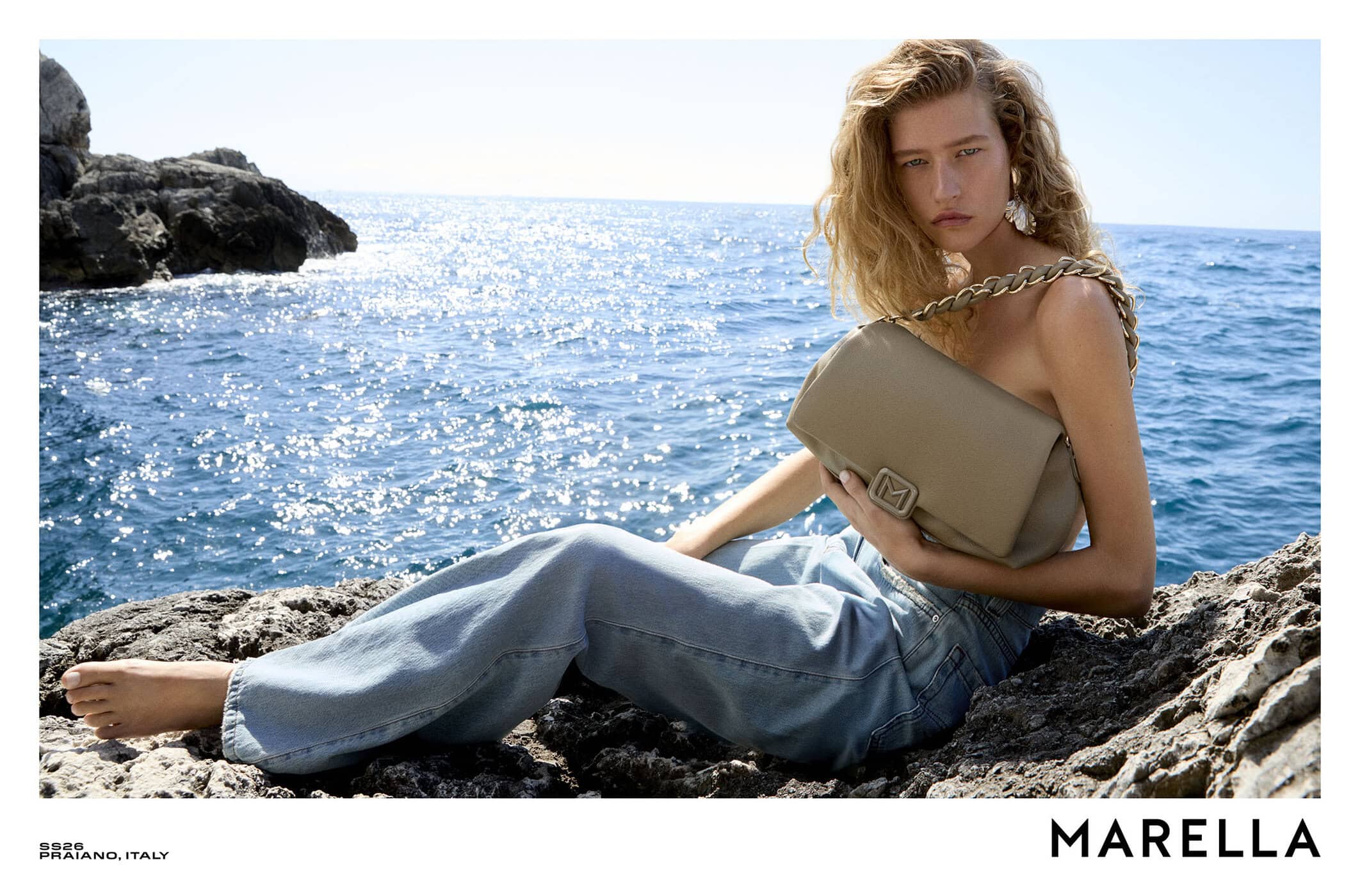 Model seated on coastal rocks wearing light denim jeans and holding a taupe leather shoulder bag against the ocean