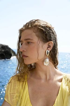 Close-up portrait of a woman with wet hair wearing a yellow dress and sculptural silver earrings by the ocean.