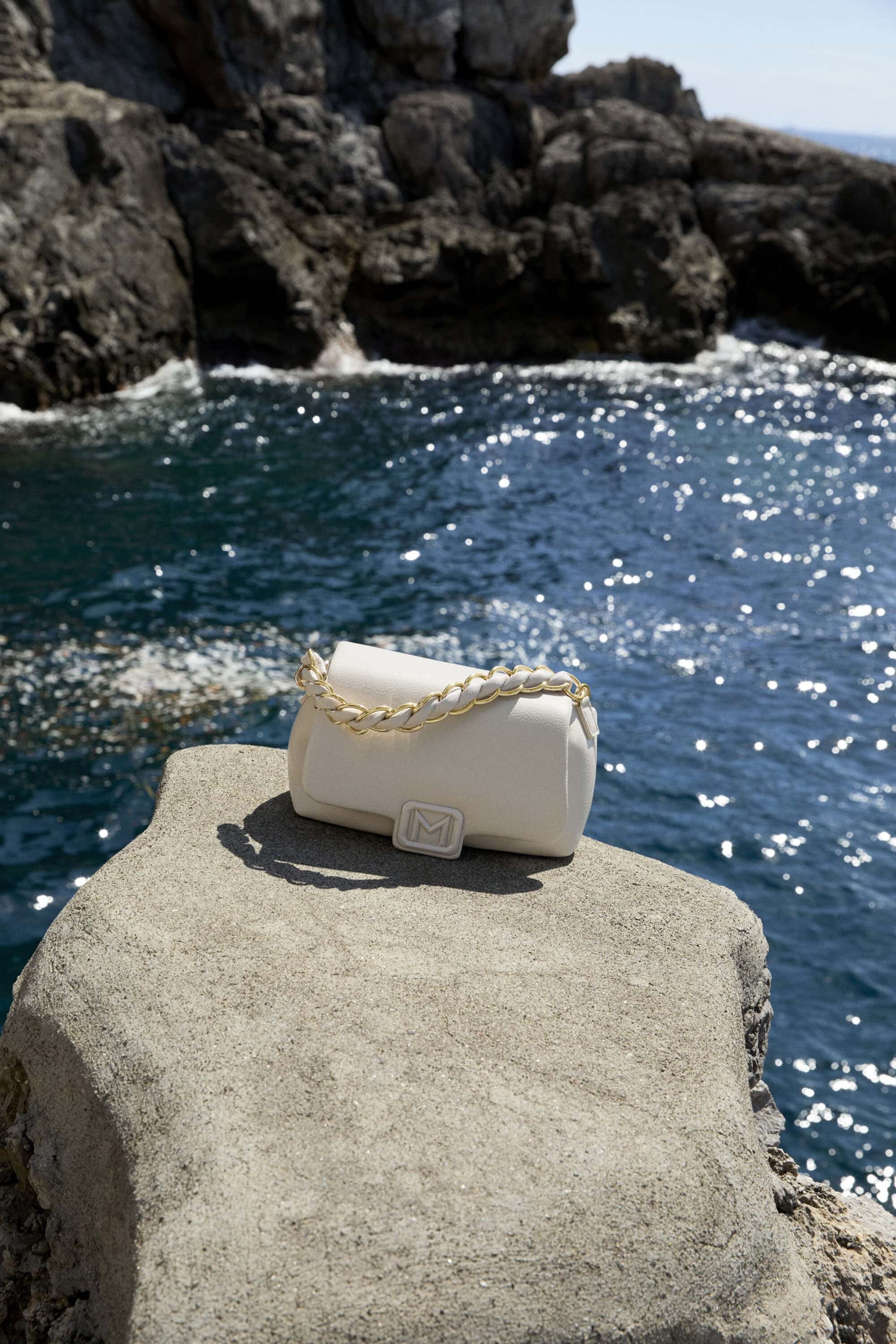White handbag with gold chain strap resting on a textured concrete ledge overlooking ocean water against a rocky background