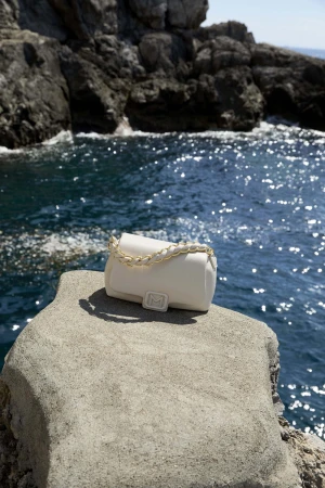 White handbag with gold chain strap resting on a textured concrete ledge overlooking ocean water against a rocky background
