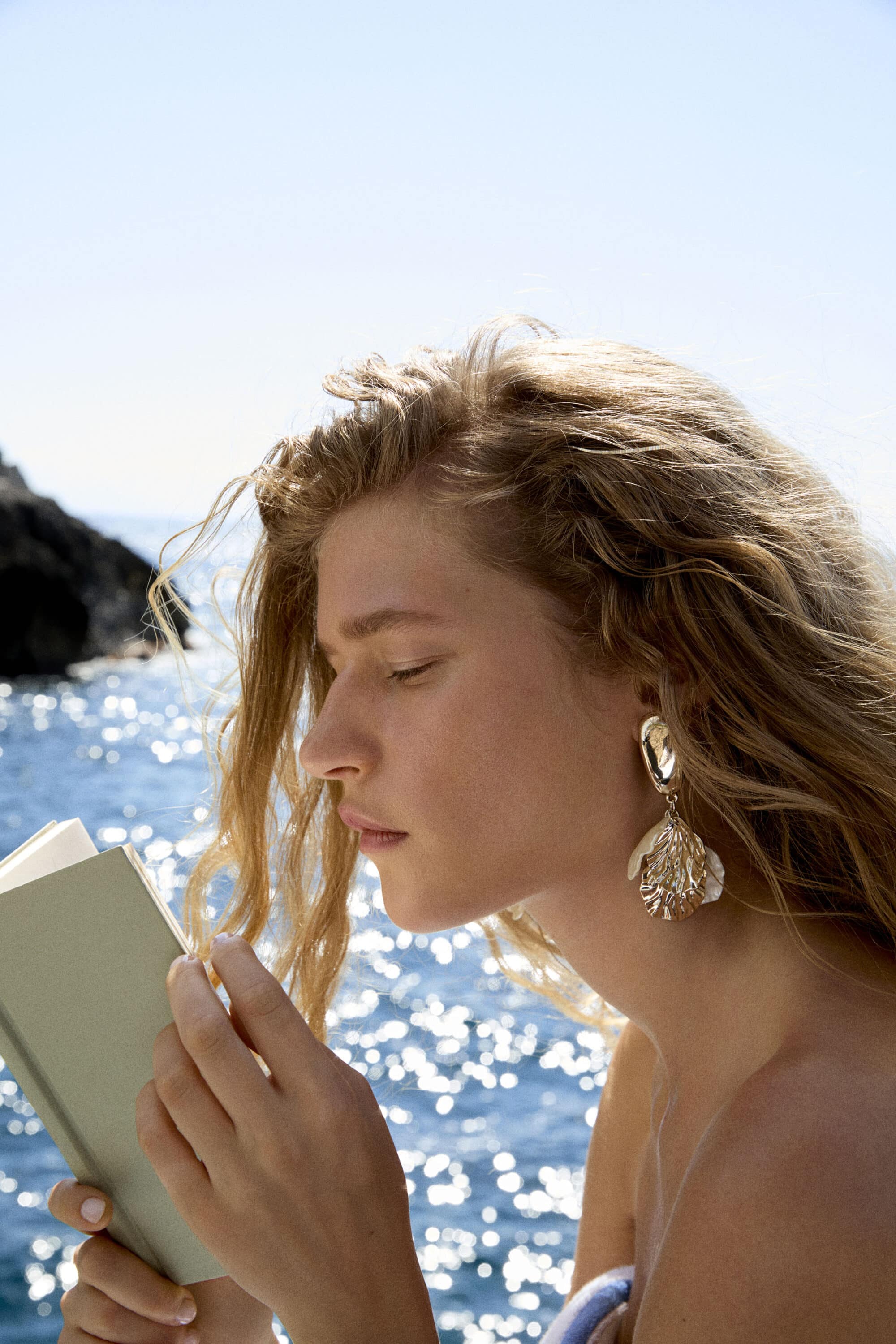 Model reading a book outdoors by the sea wearing large statement earrings for the Marella summer ad campaign.