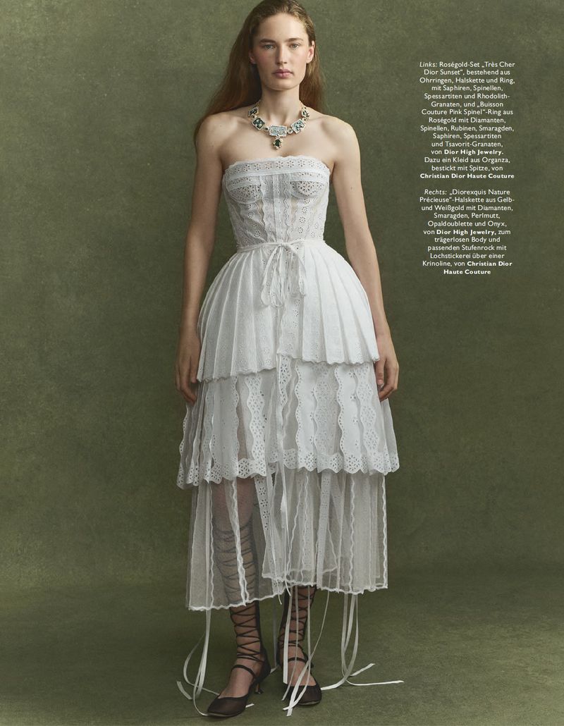 Fashion editorial shot of a woman in a white tiered dress with lace trim, complemented by a statement necklace, against a textured green backdrop.