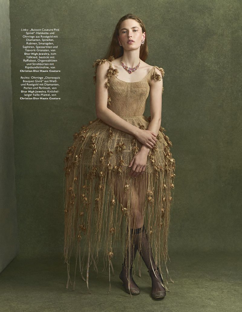 A woman in an avant-garde dress poses in a studio with textured backdrop. She wears a necklace and knee-high socks, capturing high-fashion.