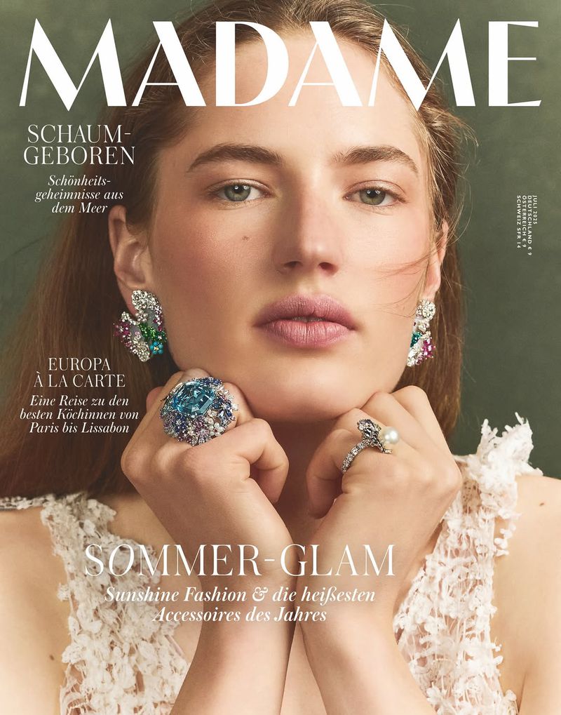 Magazine cover featuring a woman with light hair, ornate earrings, and large gemstone rings, set against a dark green background.
