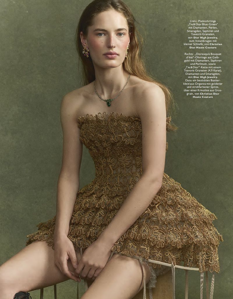 A woman with long brown hair poses in a gold lace strapless dress, with jewelry, against a mottled green studio backdrop for a fashion shot.