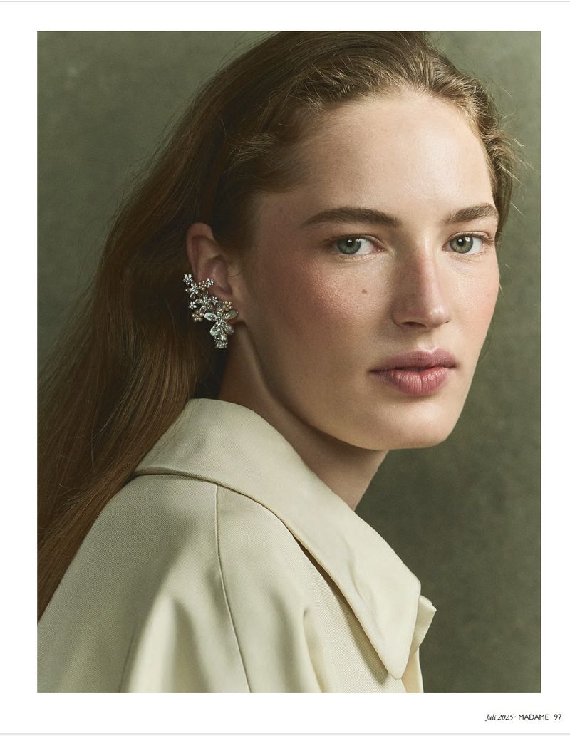 Close-up studio portrait of a young woman with long brown hair wearing an ornate earring, beige jacket, and light makeup against an olive background.