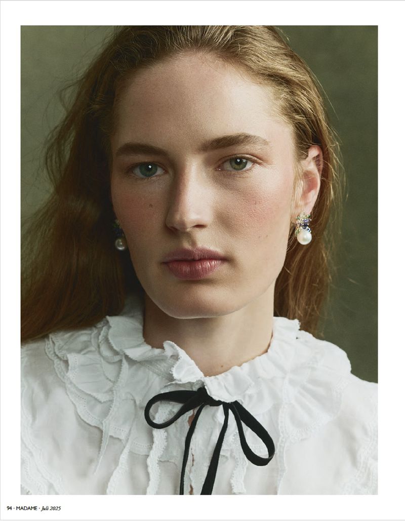 Close-up portrait of a fair-skinned adult woman with long auburn hair wearing pearl earrings and a white frilly shirt with a black ribbon.