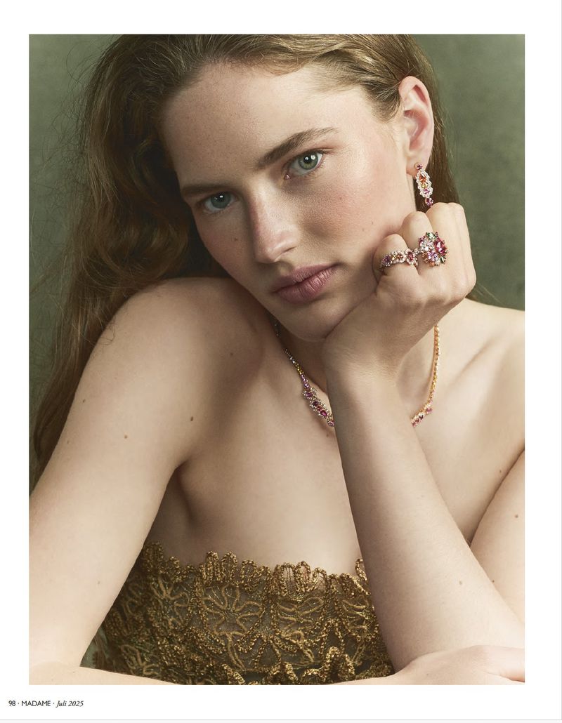 Close-up of a woman with light skin wearing gold lace top and jewelry including a ring, necklace, and earrings in a studio setting.