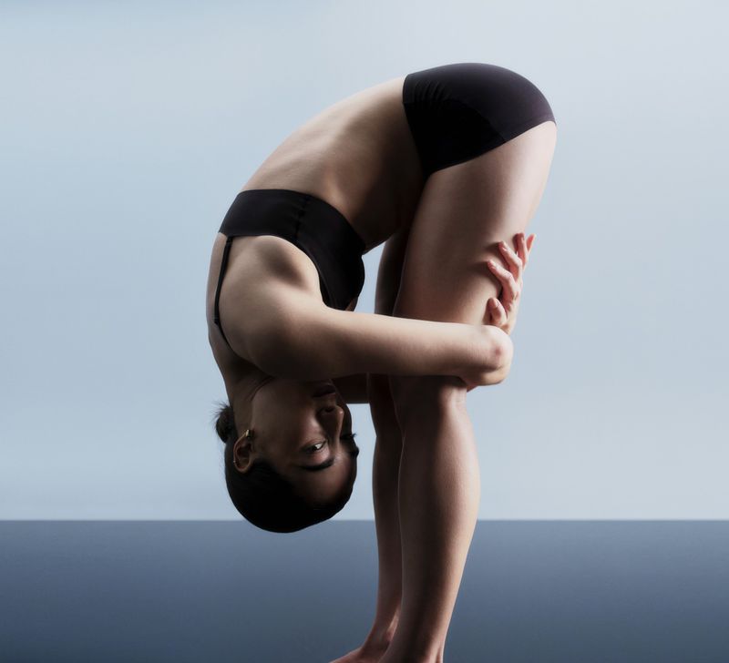 Woman in black sports bra and shorts bending forward in a yoga pose against a light blue background with dark blue at the bottom.