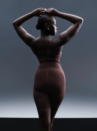 Model with braided hair wearing brown lingerie and high-waisted shorts posing against a grey studio backdrop
