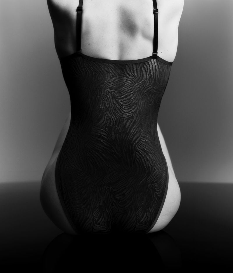 Monochrome rear view of a seated woman wearing a textured black patterned one-piece bodysuit against a grey studio background